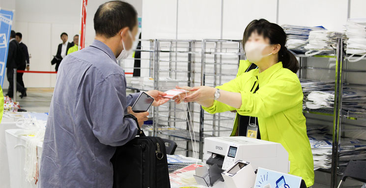 受付スタッフが来場者に印刷された赤い入場パスを渡している様子
